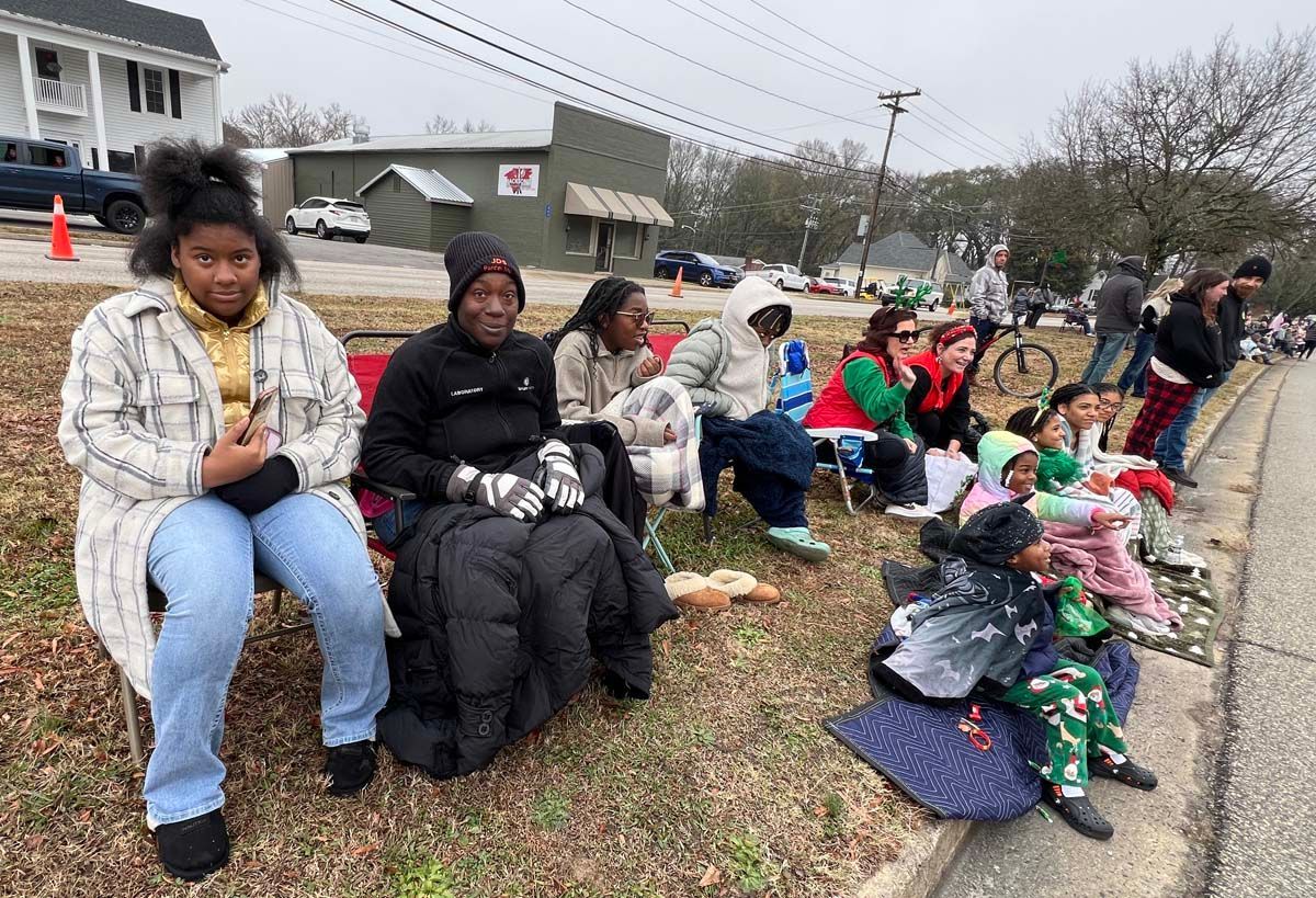 People seated along a roadside in a neighborhood, some holding holiday decorations and bags.