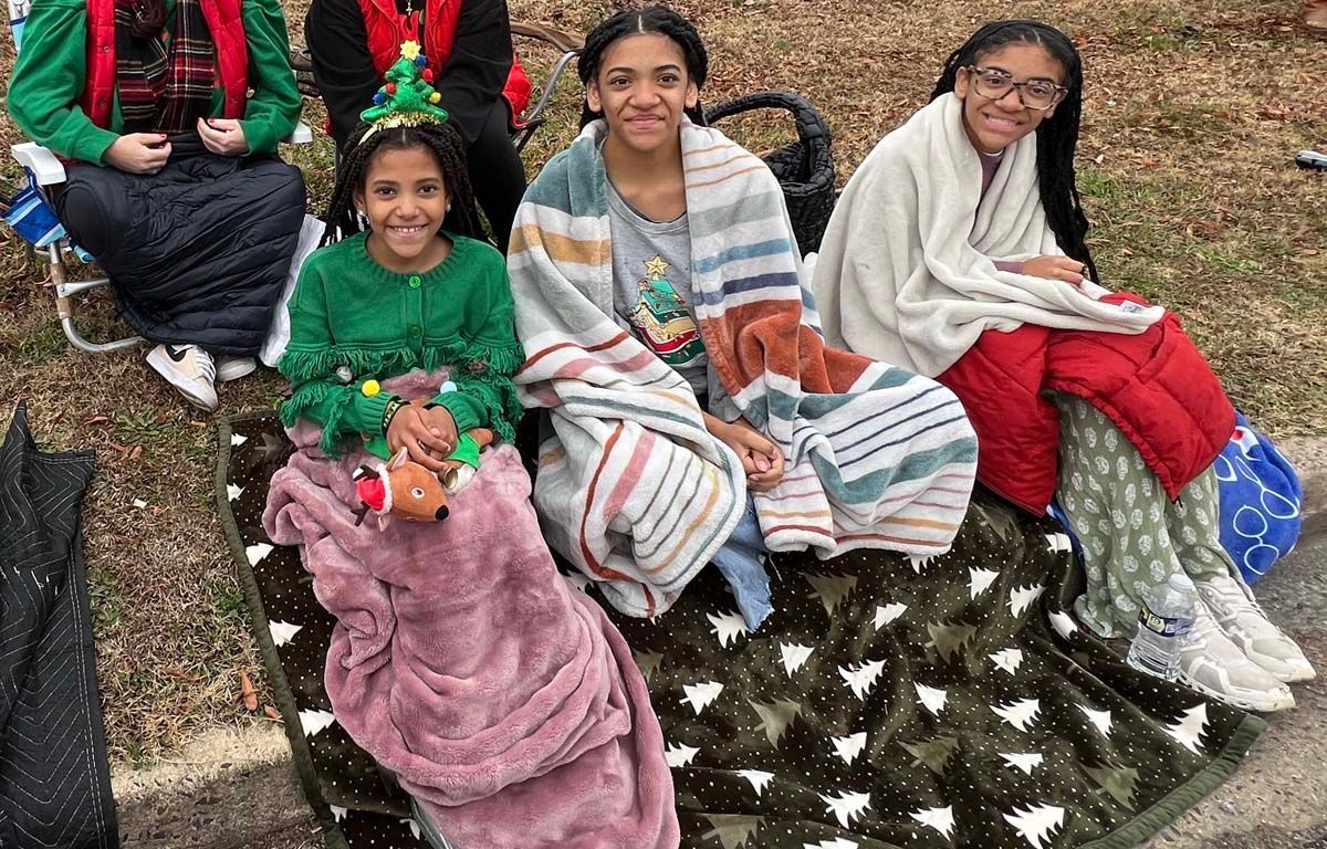 Three people sitting on a grassy slope in colorful winter blankets, smiling and posing outdoors.