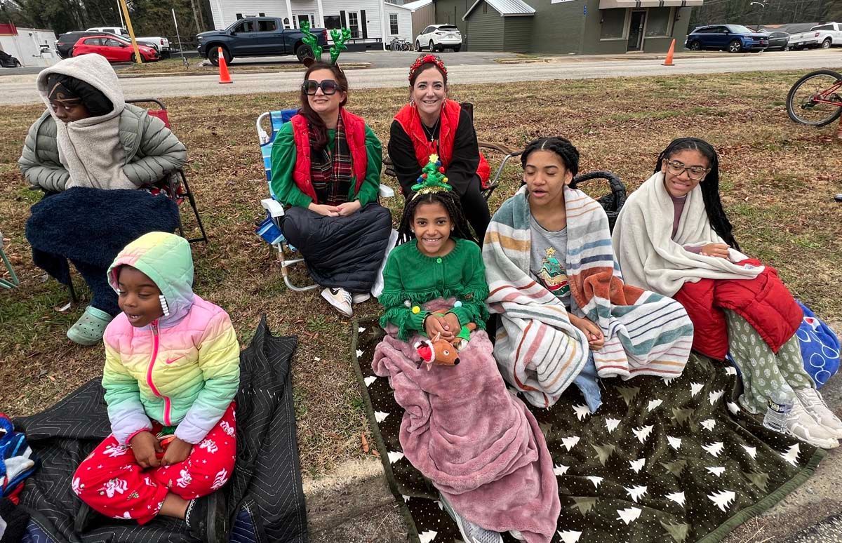 Seven people sit on a wet curb in colorful blankets during a chilly outdoor event.