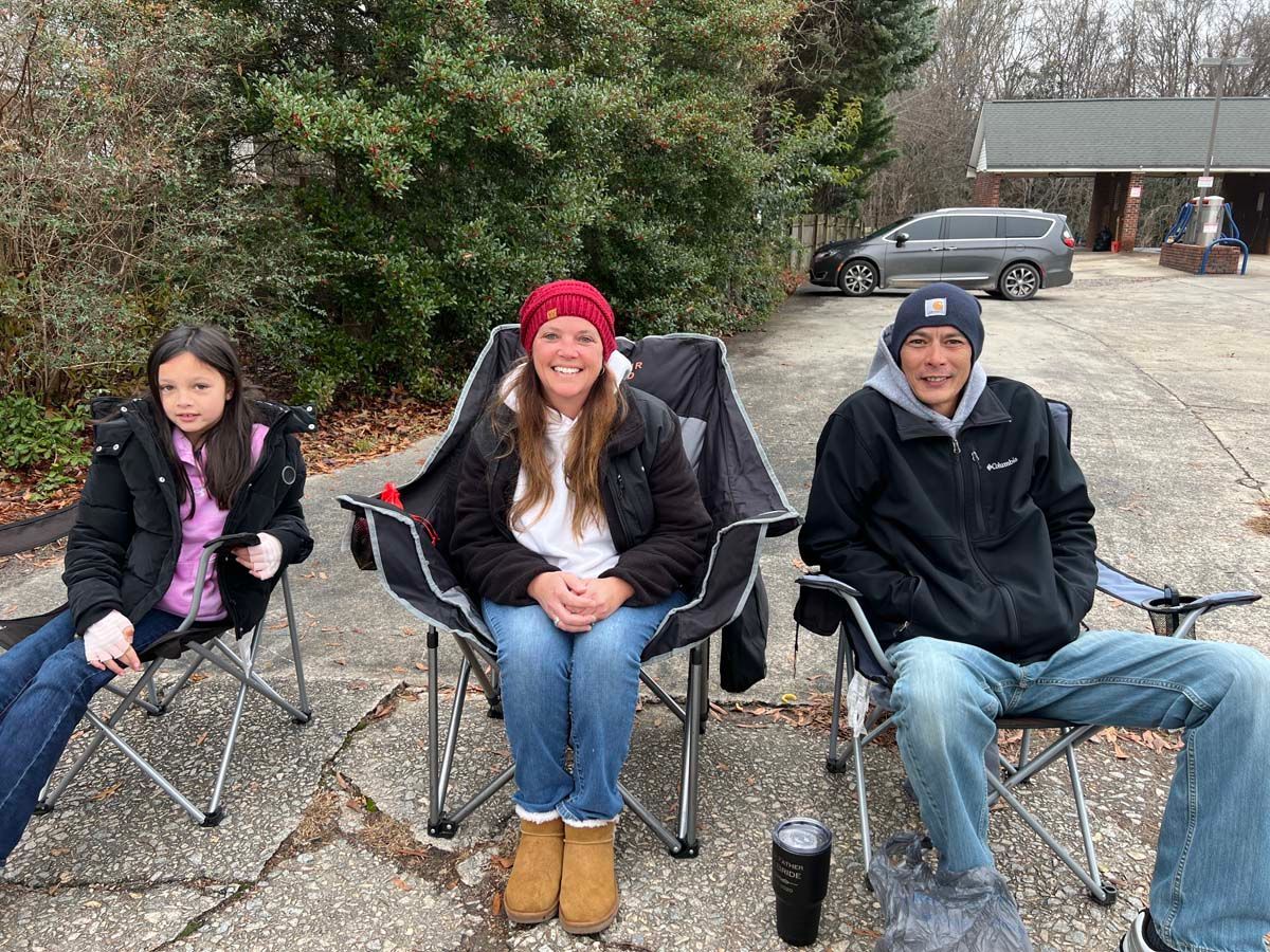 Three people sitting in folding chairs outdoors on a driveway, wearing jackets and winter hats.