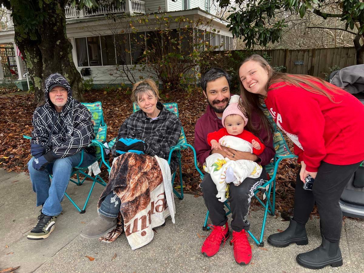 Five people seated outdoors in folding chairs, smiling at the camera, with a baby in the center.