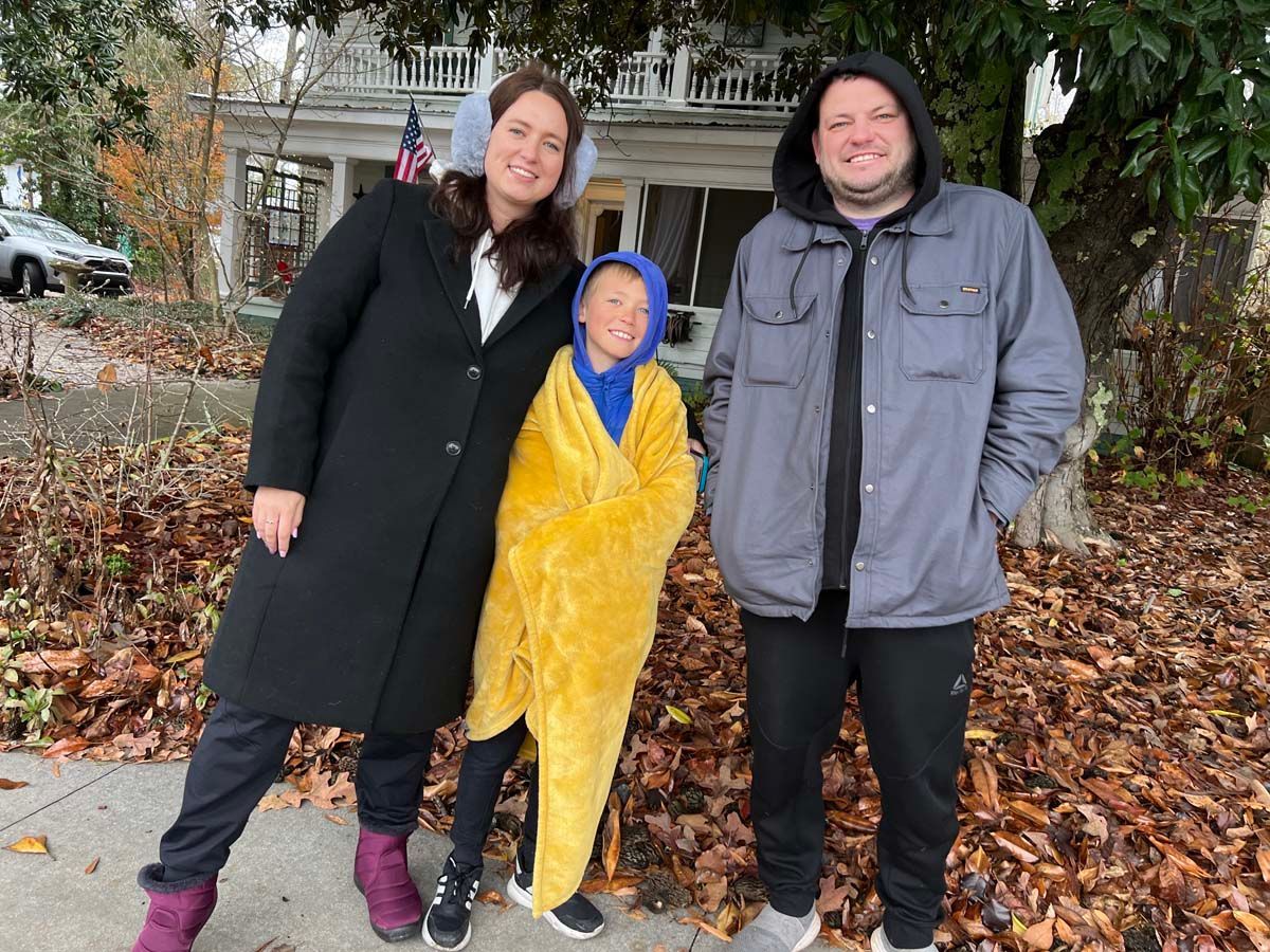 Three people standing outdoors in front of autumn leaves, one wrapped in a yellow blanket.