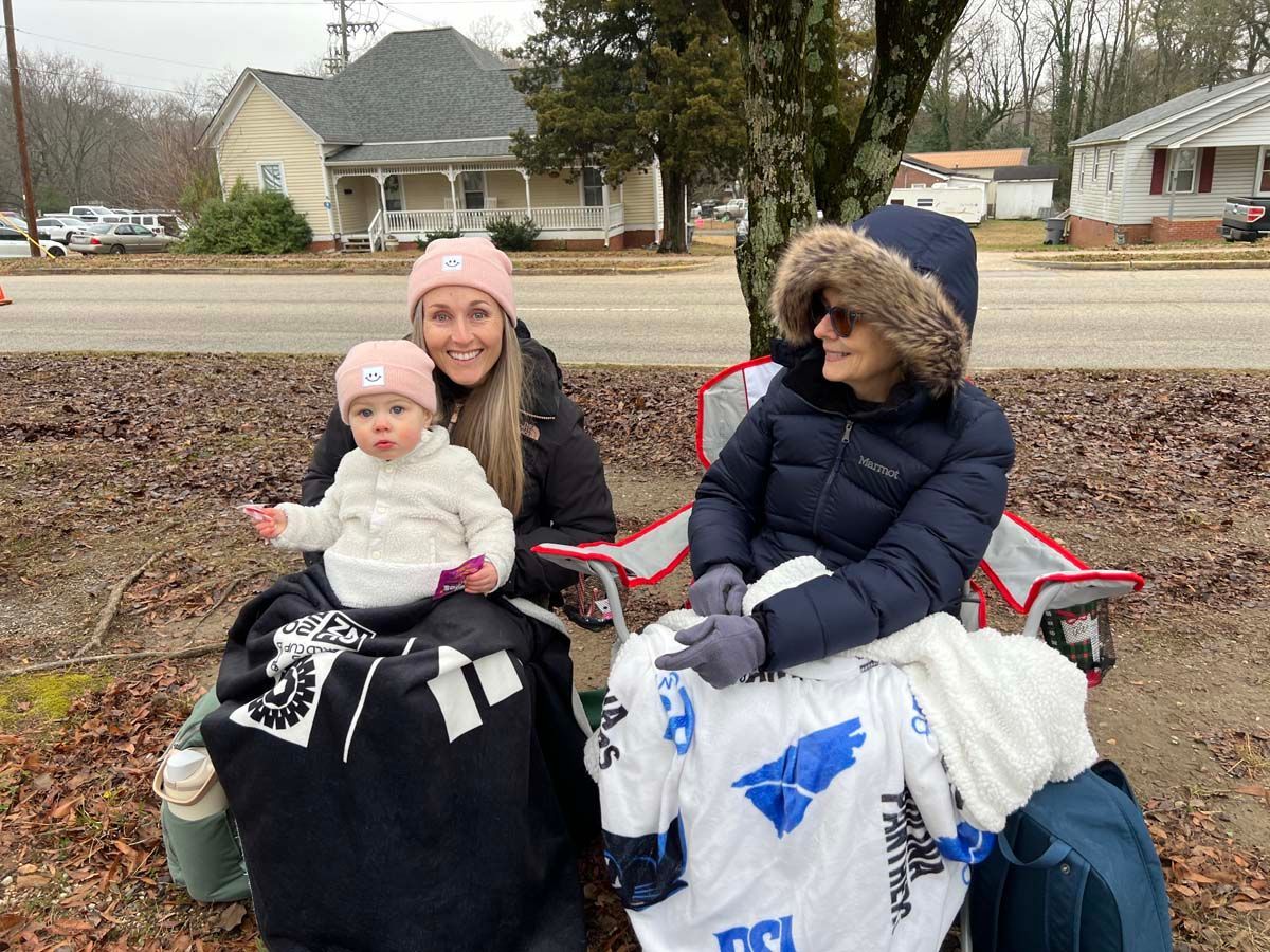 Three people seated outdoors in winter clothes, holding blankets on a chilly day.