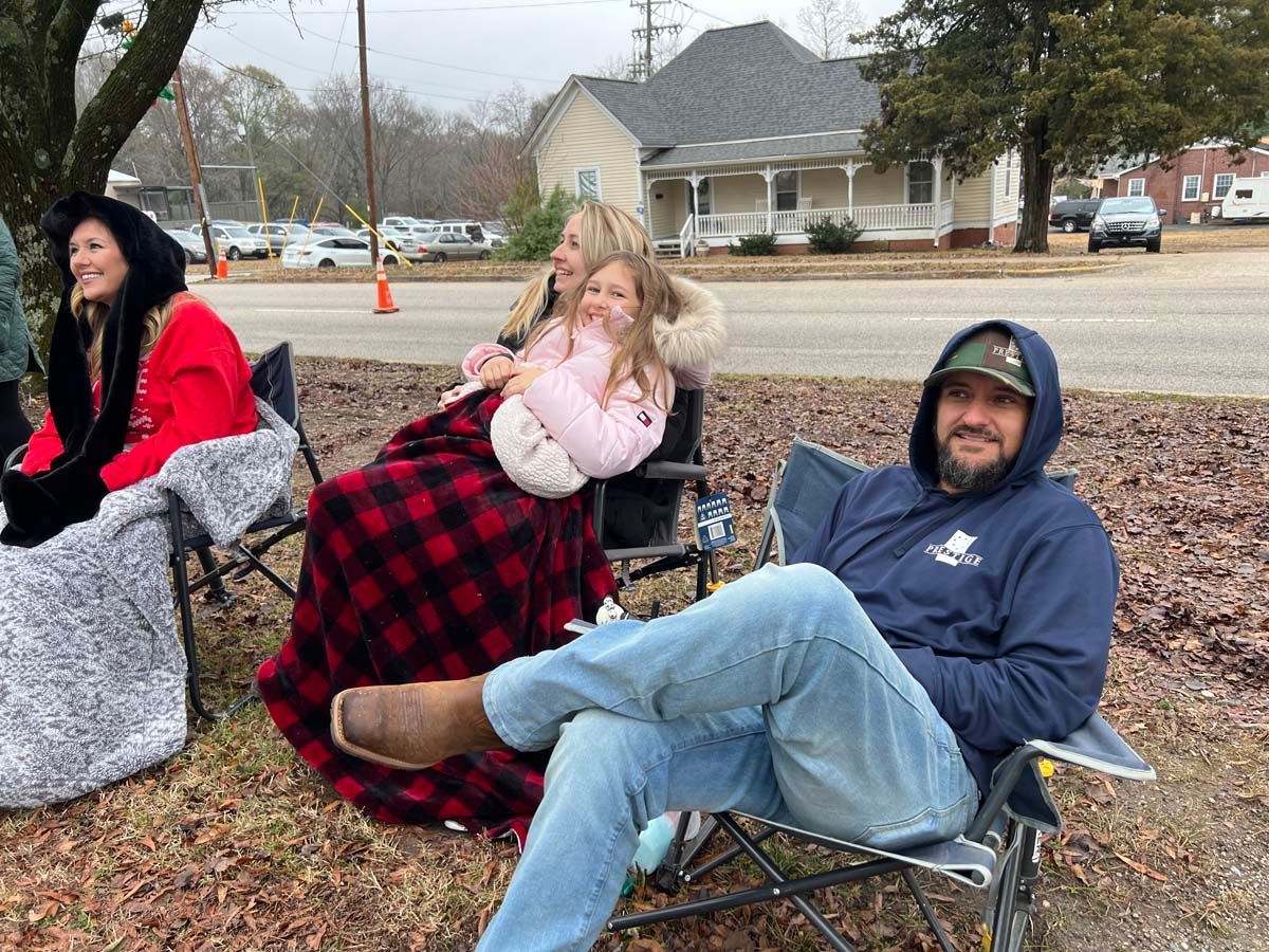 Three people sitting in camping chairs outdoors on a chilly suburban street, bundled in blankets.
