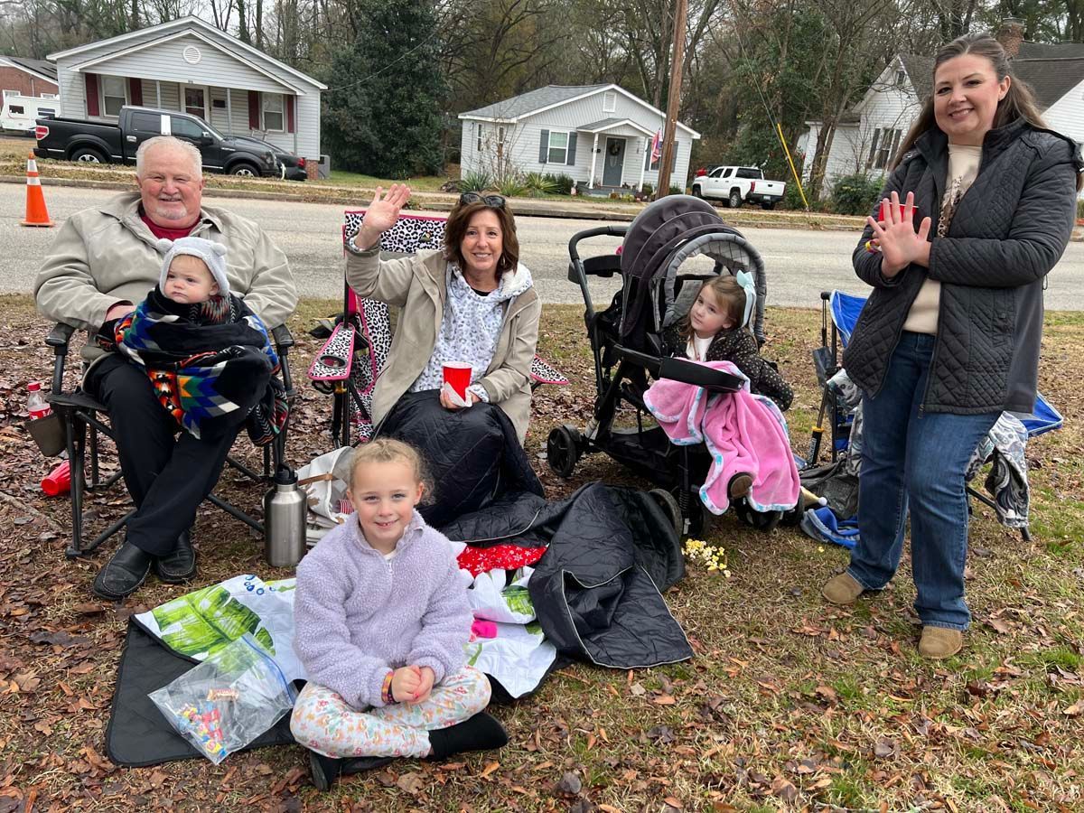 Children and adults sitting outdoors, with strollers and wrapped gifts, waving at the camera