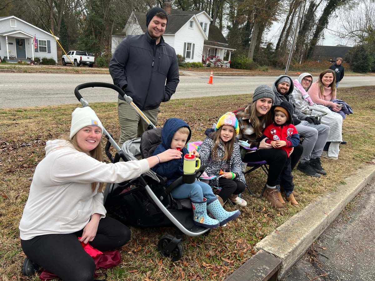 People seated outdoors along a curb, smiling and waving with a man standing behind them on a residential street.
