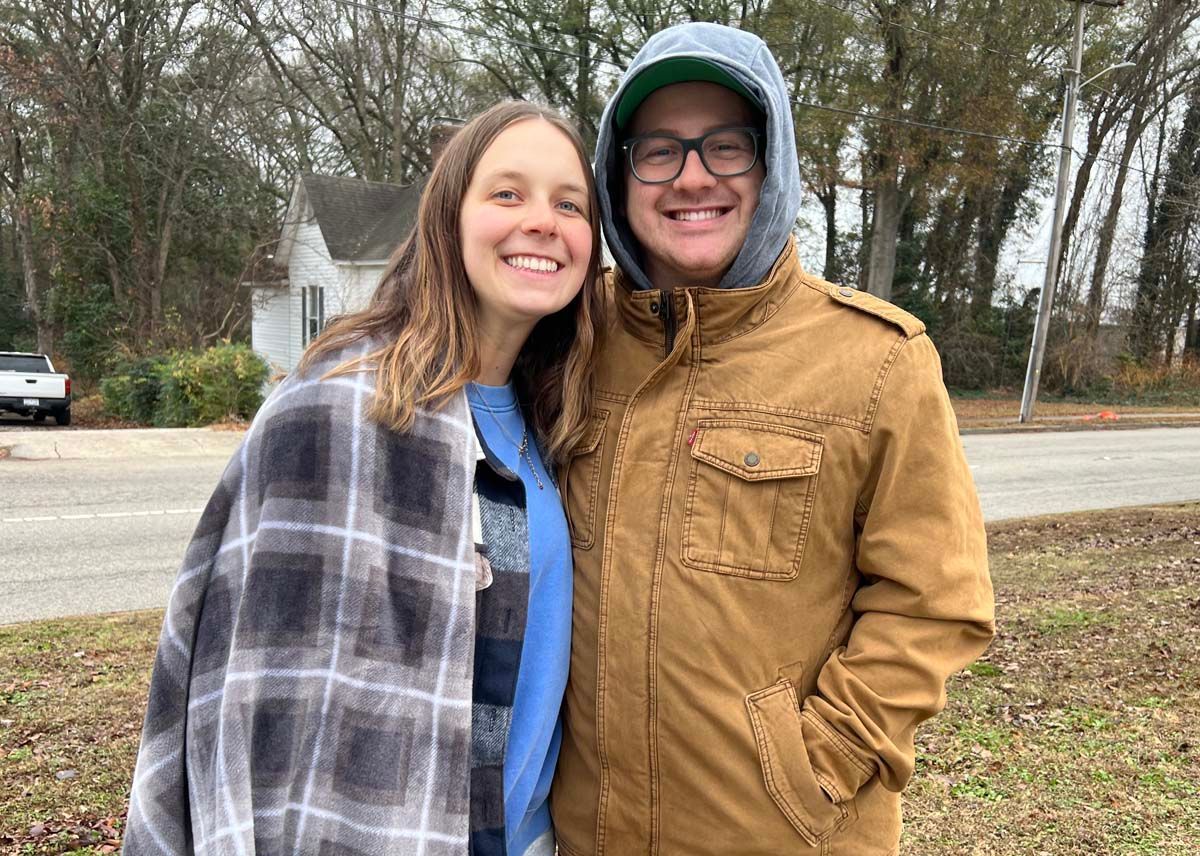 Two smiling people outdoors, one in a plaid jacket and one in a tan coat with a hood.