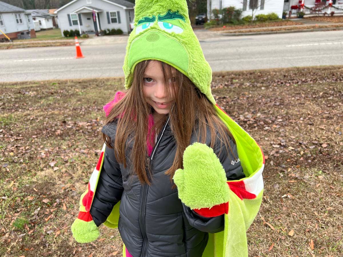 Child in a neon green monster hat and gloves standing outdoors near a road.