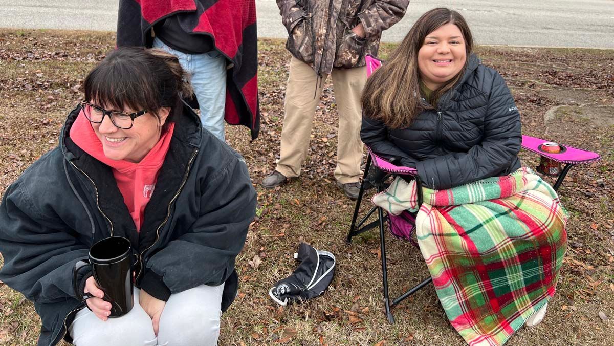 Two people sitting outdoors in cold weather, wrapped in blankets and smiling near bare trees.