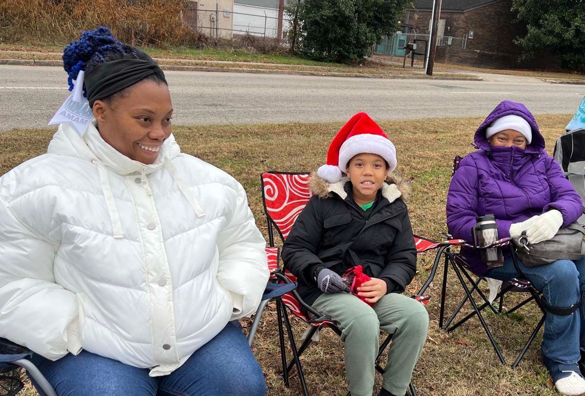 Three people sitting outdoors in winter coats, one wearing a Santa hat and smiling, on folding chairs.