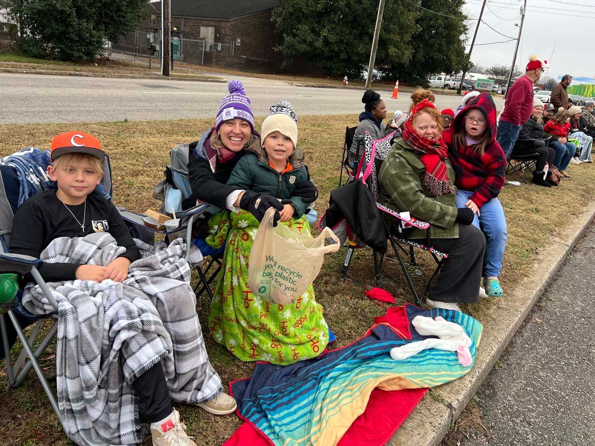 People in winter coats sit on a curb, smiling, holding blankets and bags during a cold-weather event.