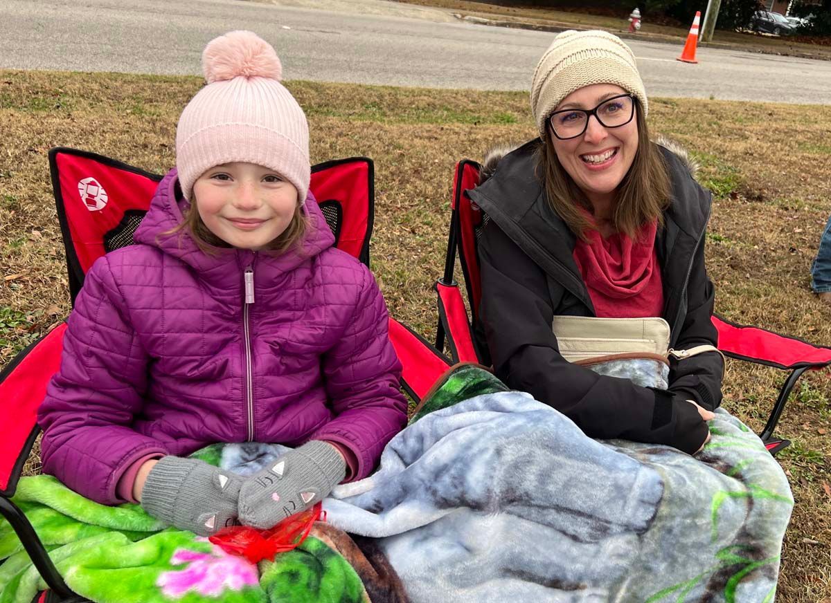 Two people sitting in red chairs outdoors, bundled in winter clothes and blankets, smiling at the camera