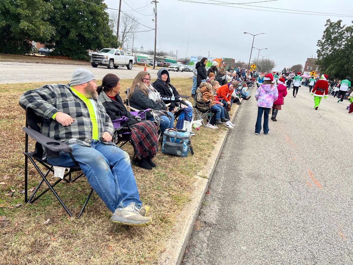 People seated in folding chairs along a roadside, watching a parade pass by.