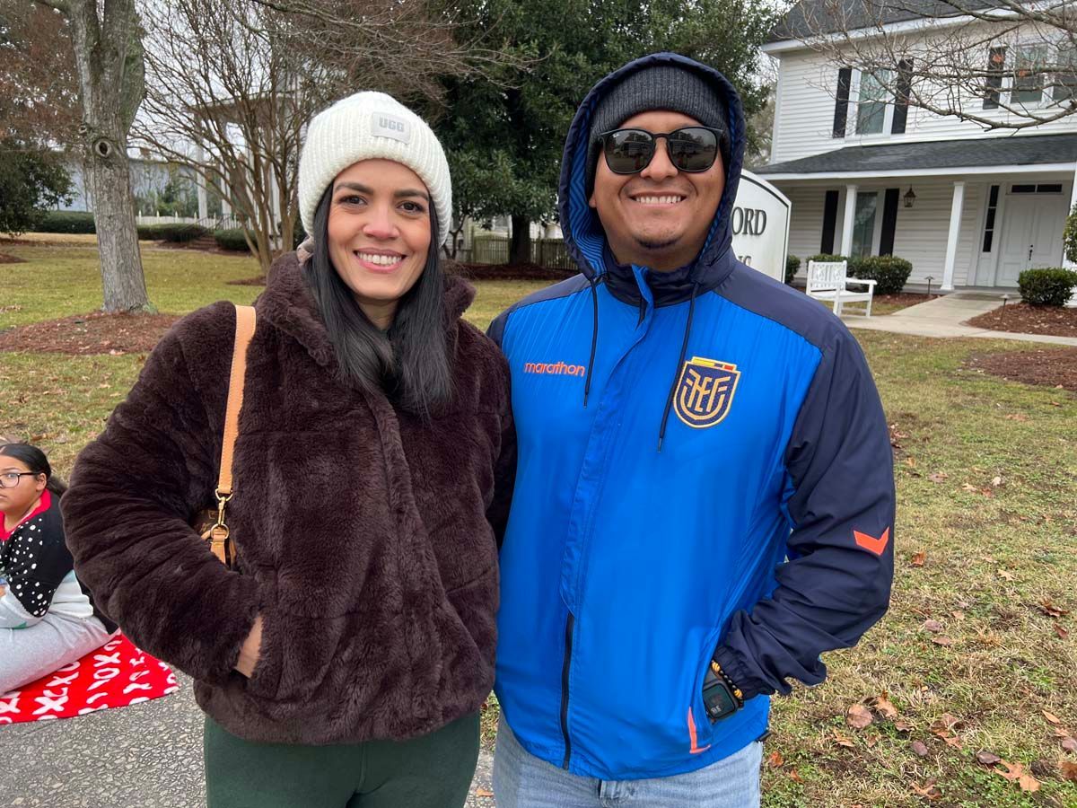 Two people smiling outdoors in winter coats and hats on a grassy lawn with houses in the background