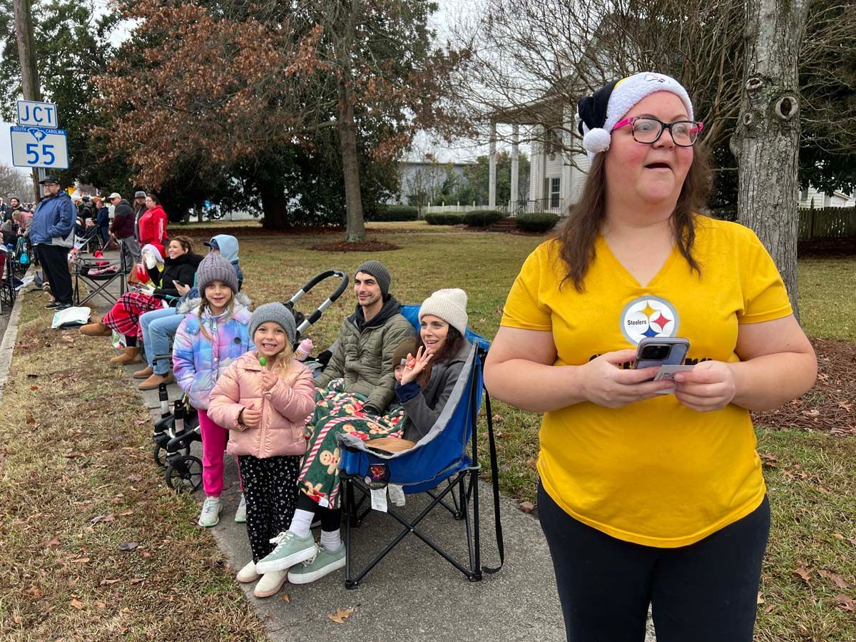 Woman in yellow shirt holding a trophy at a sidewalk parade, with children seated along the curb.