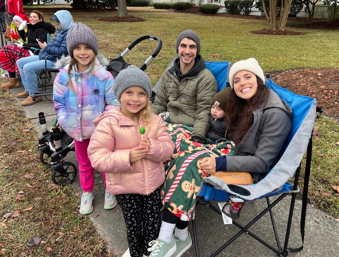 Four smiling children and two adults sit outdoors in folding chairs on a chilly day.