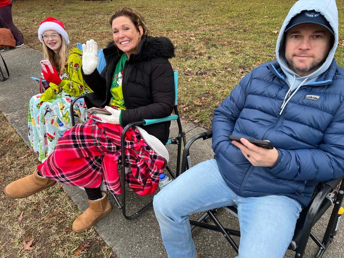 Three people sitting outdoors on folding chairs, smiling, one holding a phone and another in a Santa hat.