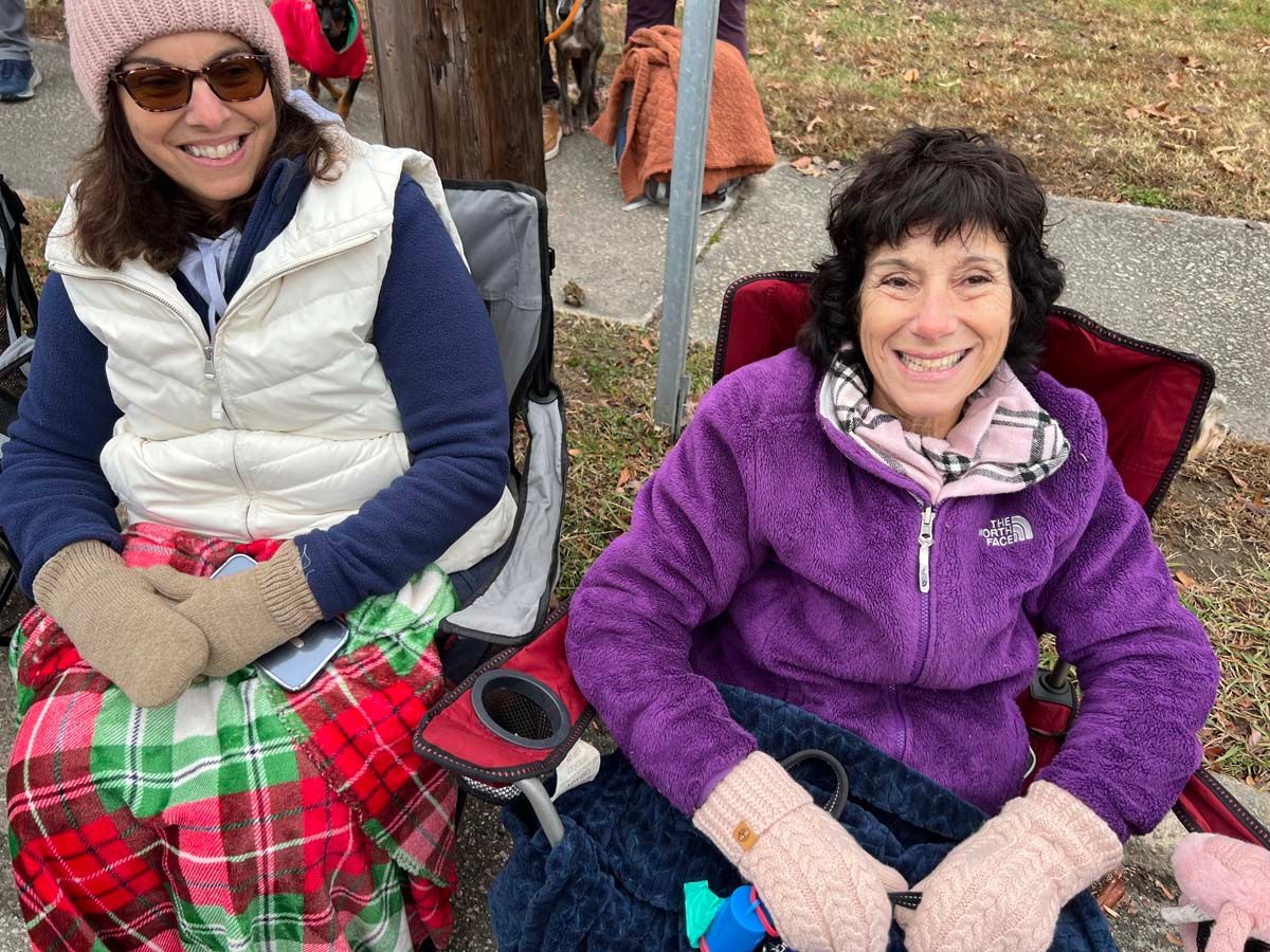 Two women in folding chairs outdoors, wearing winter jackets and smiling at the camera.