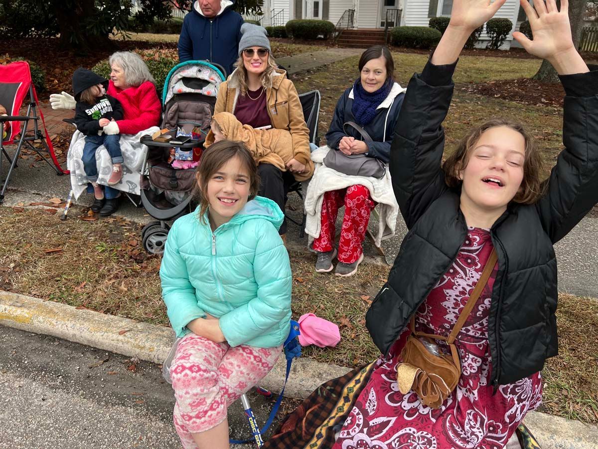 Children sitting on a curb outdoors, one girl smiling and another raising her hands, with strollers behind them