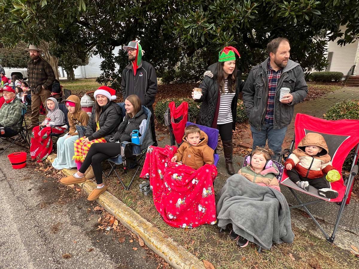 Children in festive hats sitting in red chairs along a curb under a tree