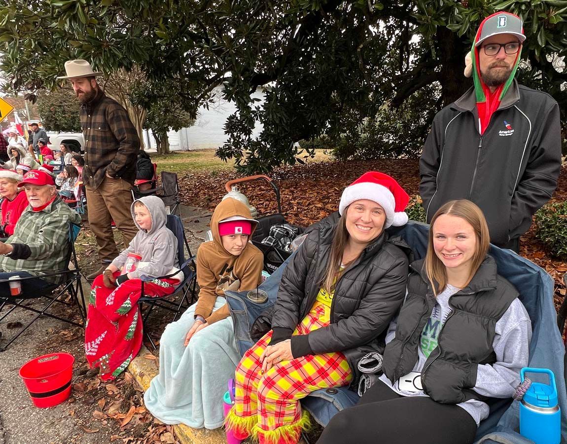 People sitting outdoors in festive winter clothes, some wearing Santa hats, smiling on a blanket-lined bench.