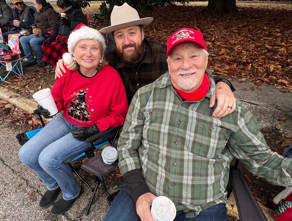 Three people in red and plaid outfits sit together outdoors, smiling with drinks.
