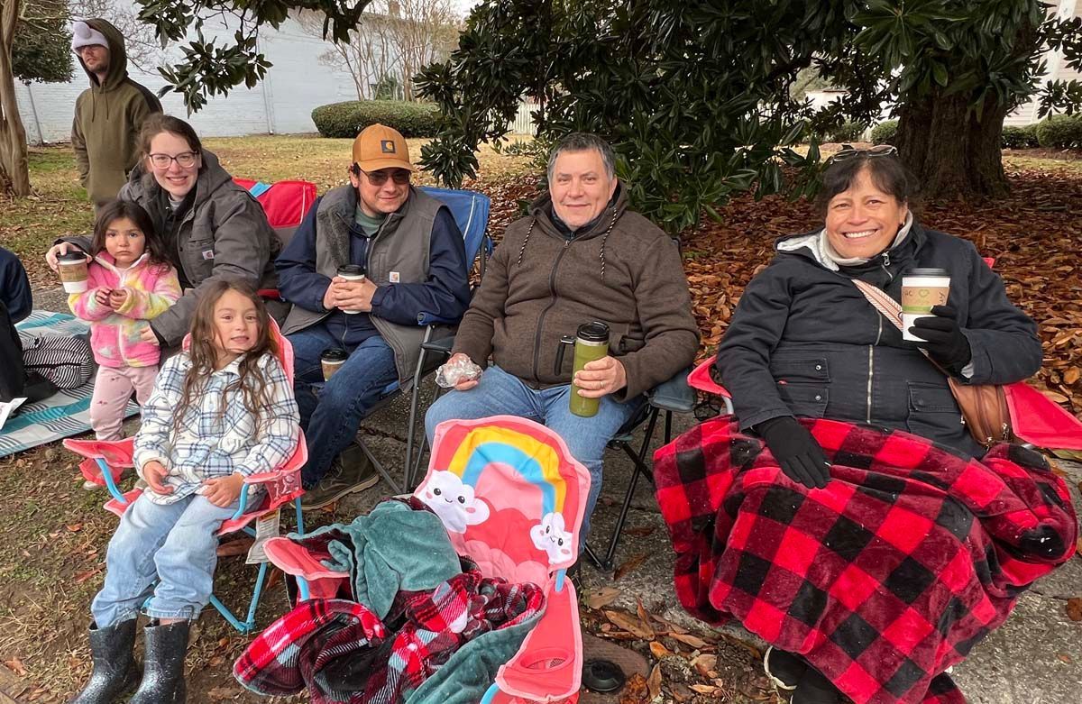 People seated outdoors on chairs and blankets, bundled in winter clothing, smiling at a gathering