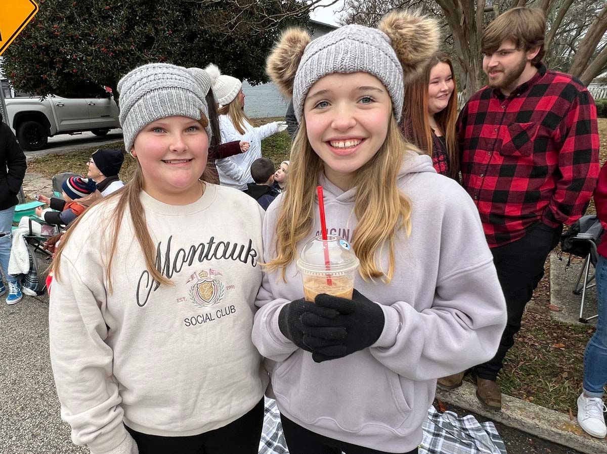 Two smiling people in winter hats and gloves standing outdoors with other people in the background