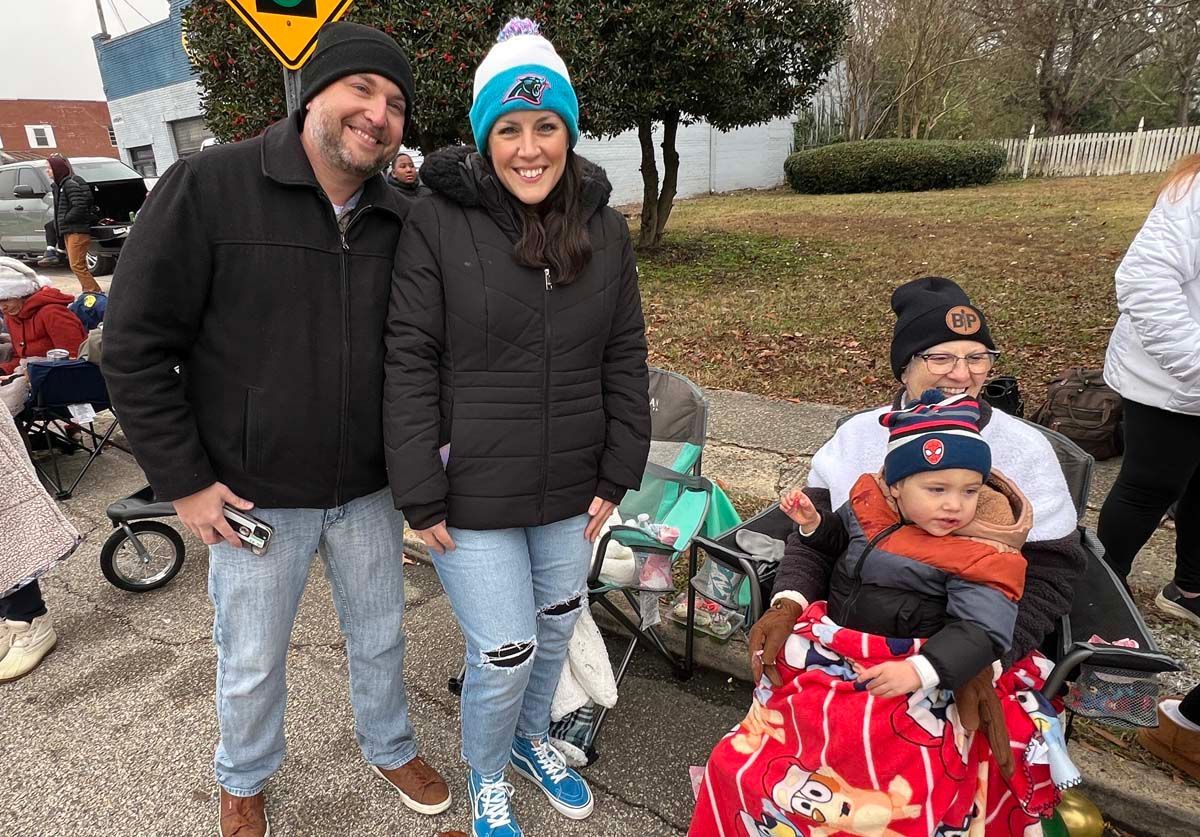 Two adults and two children smiling outdoors, with strollers and a red wagon on a cloudy day