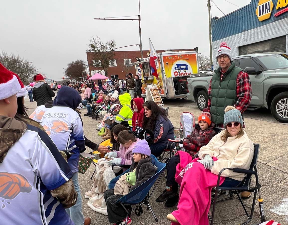 Crowd in folding chairs along a street, bundled in winter clothes, watching a parade in light snow