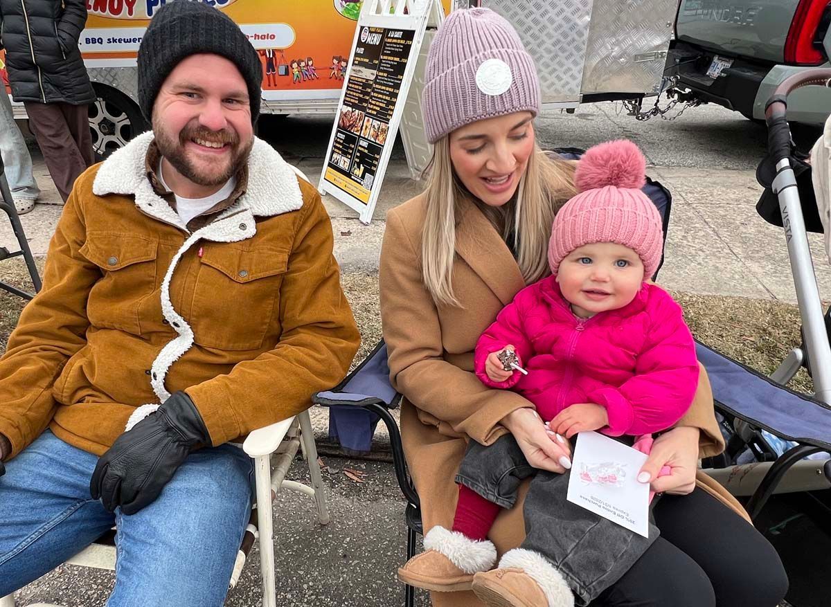 Two adults and a baby seated outdoors in winter clothes, smiling near a market stall