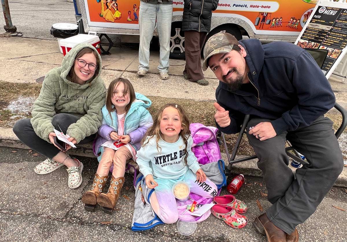 Family posing on a snowy street with two children, colorful toys, and a pet cat by an orange truck