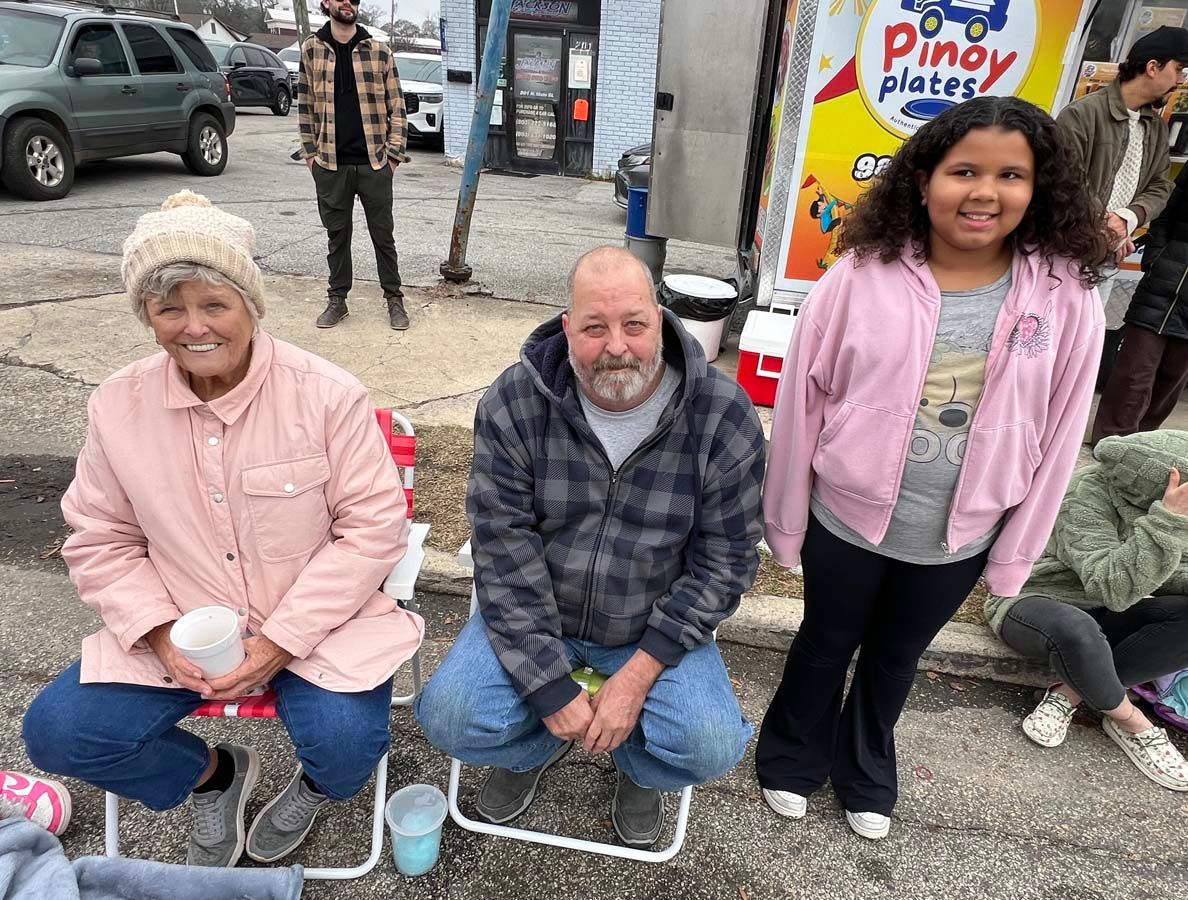 People seated outdoors on folding chairs beside a food truck, smiling at the camera.