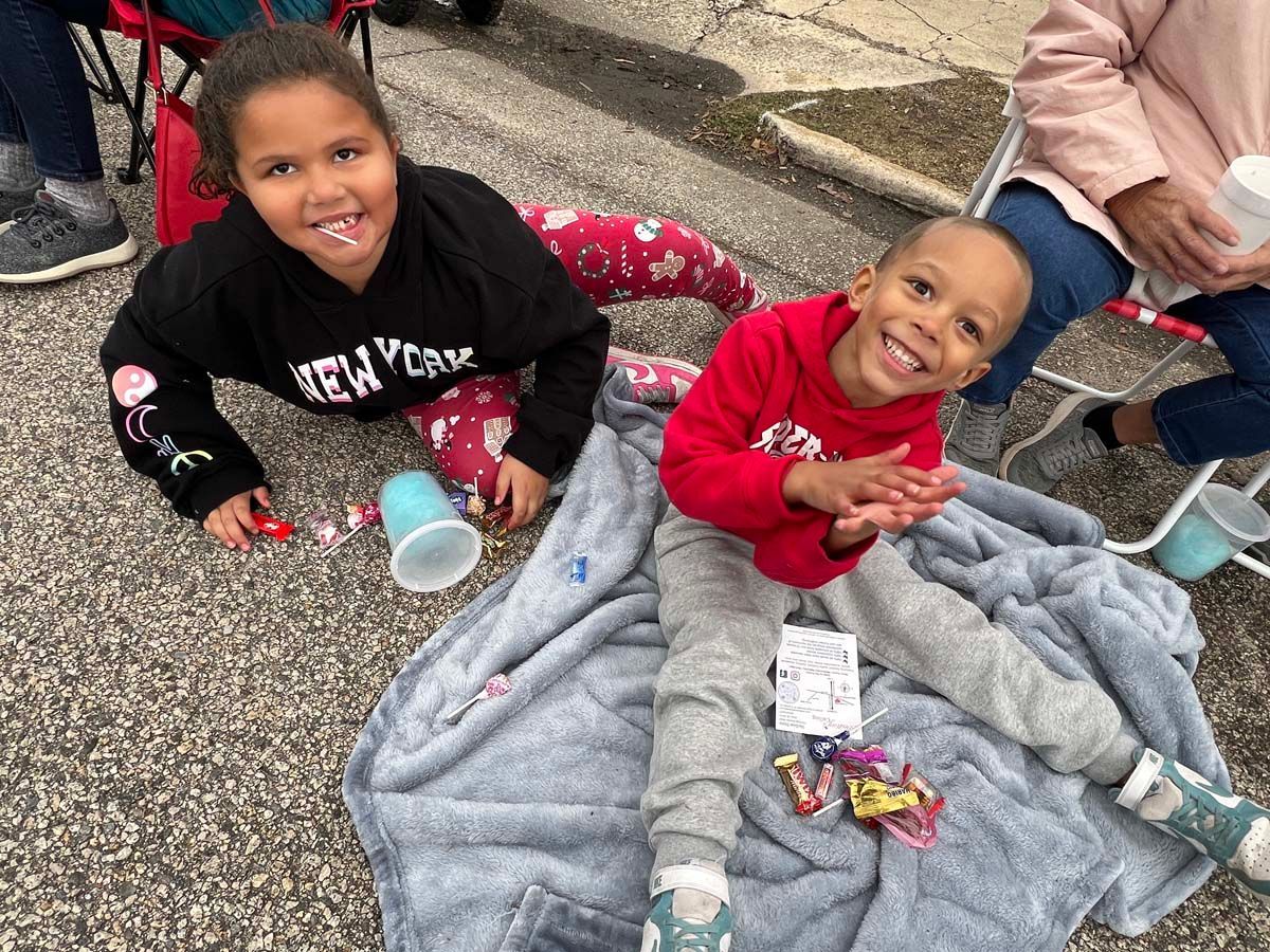 Two children sitting on pavement, smiling beside a gray blanket and snacks outdoors.