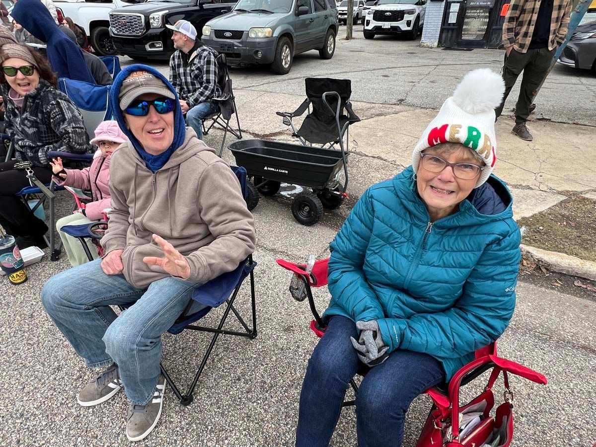 Two smiling people in folding chairs outdoors, bundled in jackets and hats, at a curbside gathering