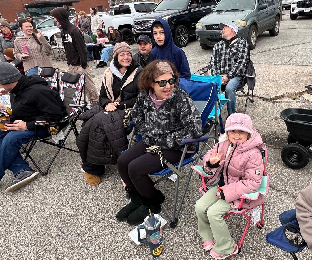 People seated in folding chairs outdoors at a casual gathering, with a child in a pink coat waving.