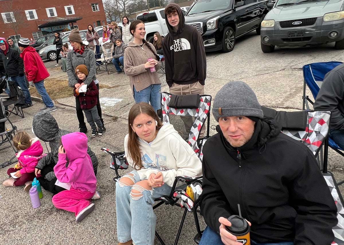People seated outdoors at an event, wearing coats on a chilly day, with others and parked cars in the background