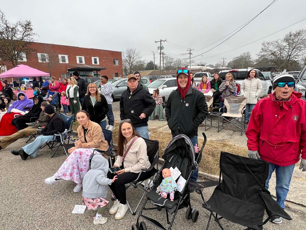 Crowd seated outdoors at a community event, with strollers, folding chairs, and a brick building in the background