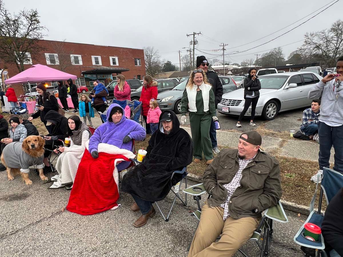 People seated outdoors at a community event, some in costumes, with cars and a brick building behind them