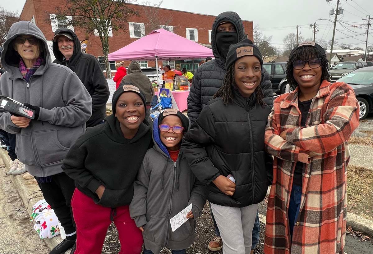 Group of smiling people posing outdoors in winter coats at a community event with tents and a brick building.