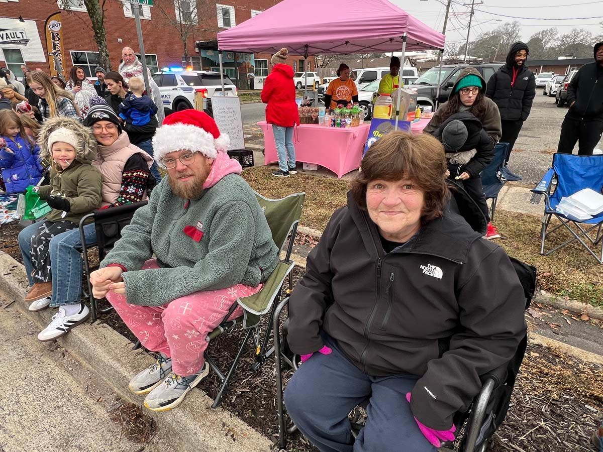 People seated outdoors at a community event under a pink canopy, smiling and bundled in jackets
