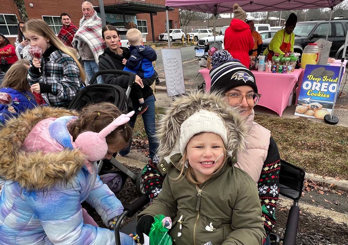 Children in winter coats at an outdoor holiday event, with a decorated table and festive crowd in the background
