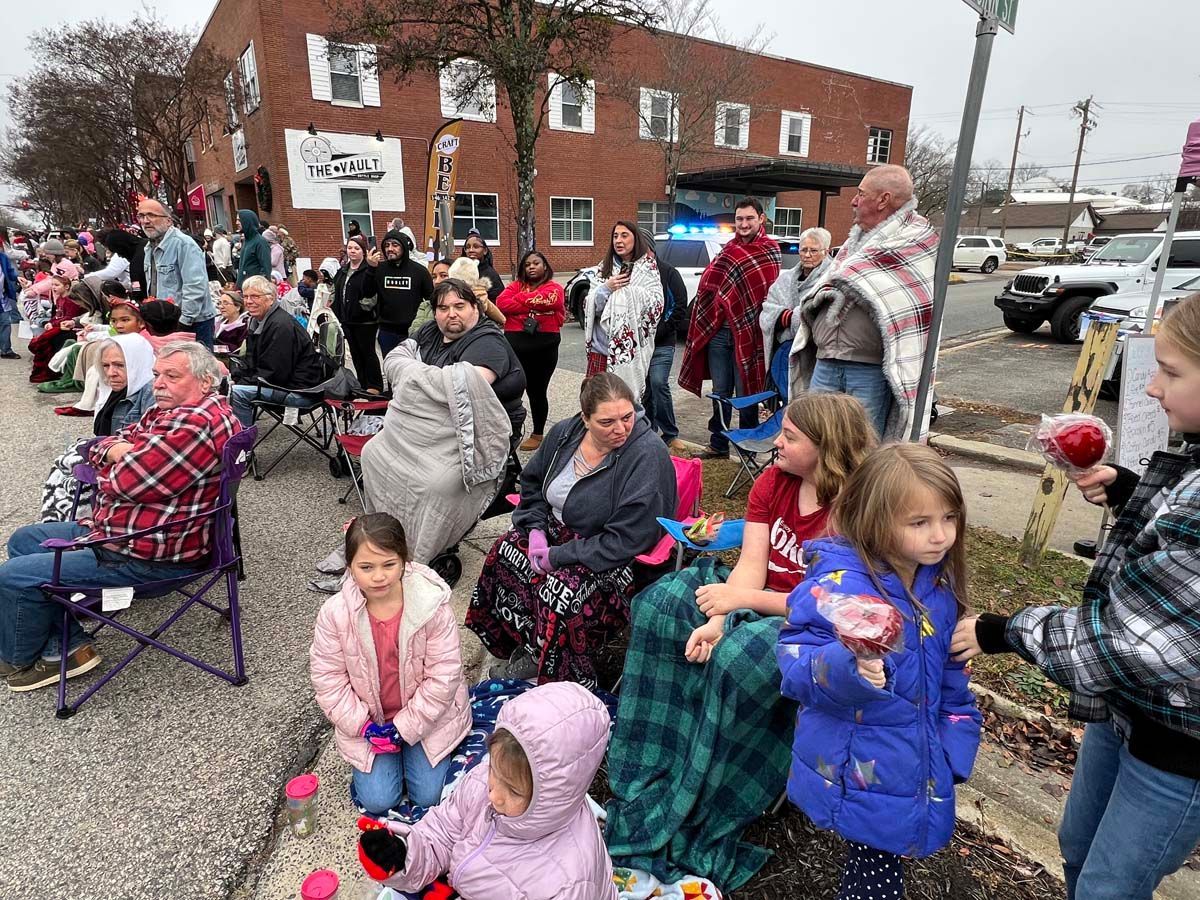 Crowd seated outdoors near a brick building, bundled in coats and blankets on a cold day.