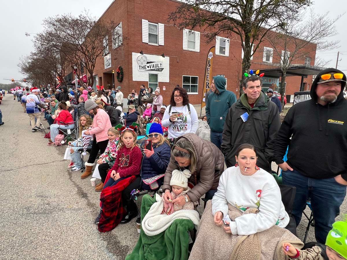 Crowd seated along a street outside a brick building during a parade or community event