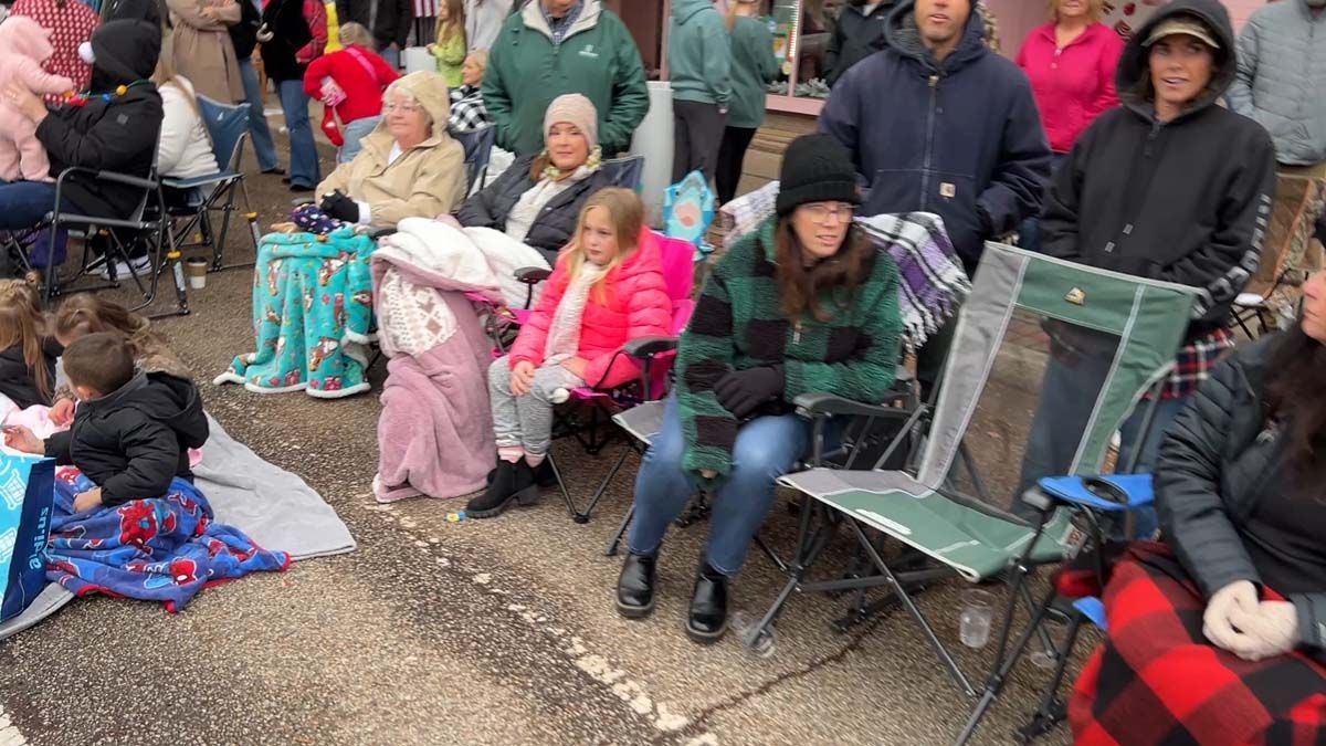 People seated outdoors on gravel, bundled in coats and blankets, watching an event.