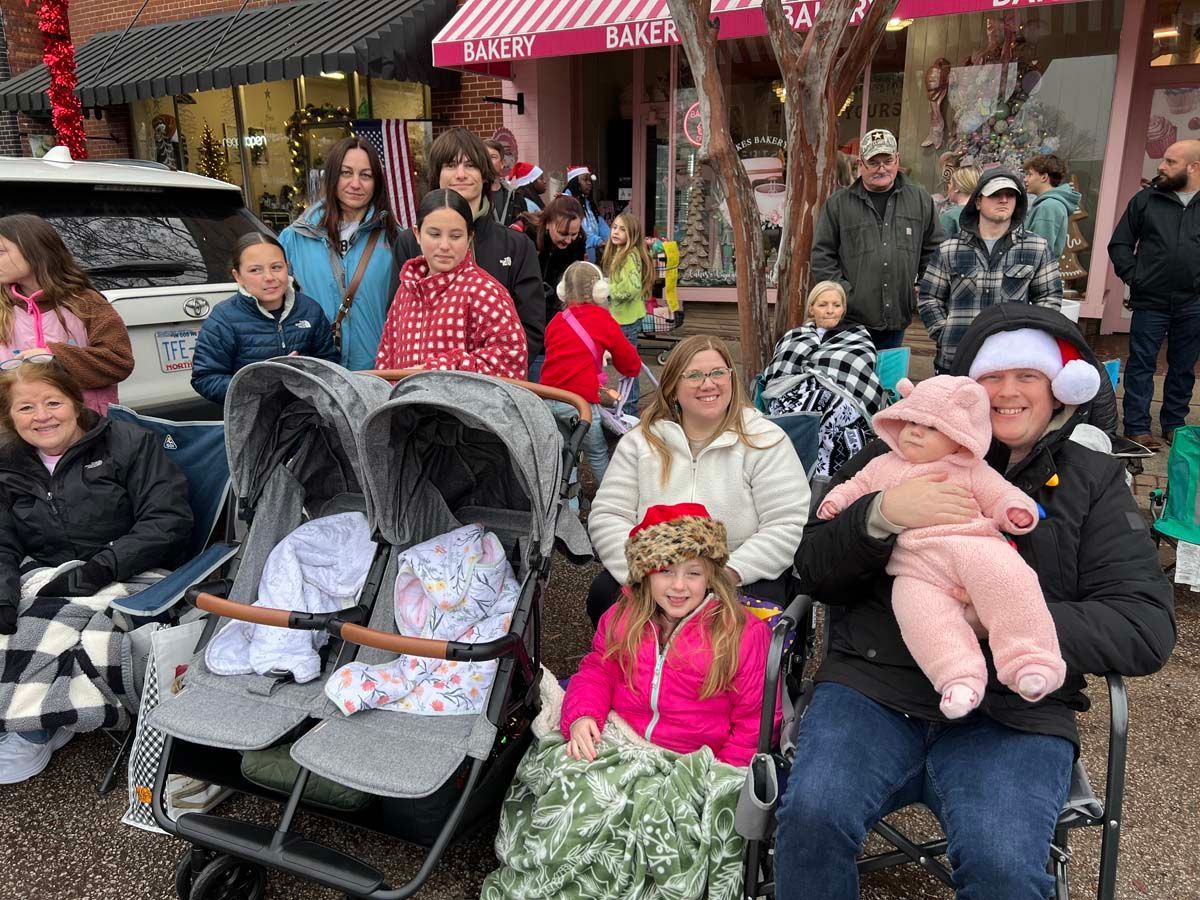 Crowd gathered outdoors near a bakery, with two strollers in front and children in festive clothing.