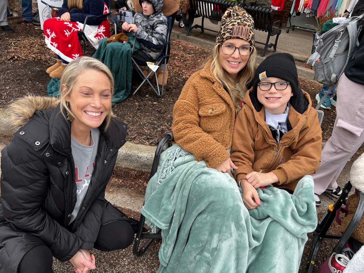 Three people smiling at an outdoor market, two seated under a mint blanket and one crouching beside them.