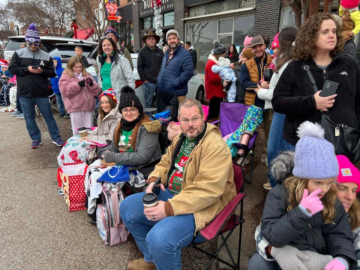 People seated along a city sidewalk, many wearing hats and coats, watching or waiting near storefronts.