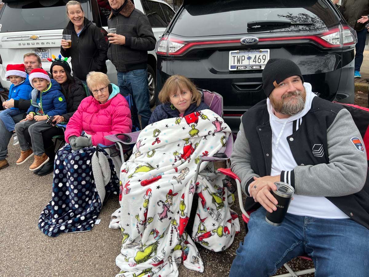 People sitting outdoors on chairs and blankets in front of a parked car, bundled up in cool weather.