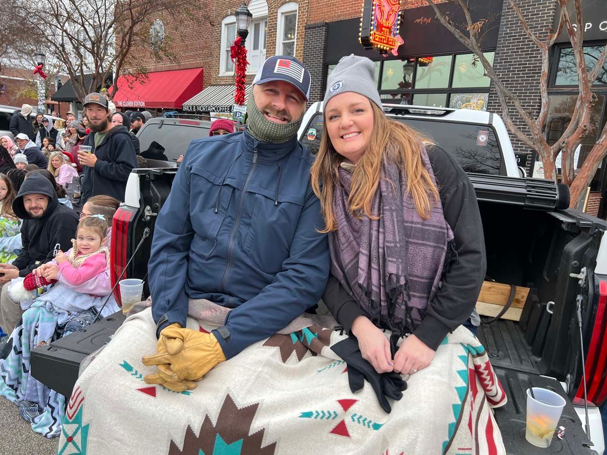 Couple sitting on a decorated pickup truck at a winter parade, smiling under a blanket